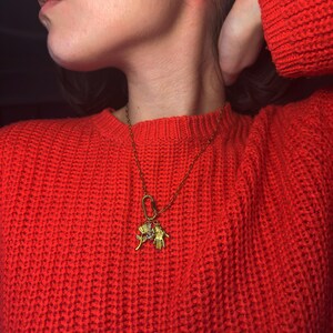 May include: A gold-colored necklace with a charm featuring a hand and a rose. The necklace is worn over a red, chunky knit sweater. The necklace has a large clasp and a small charm with a gemstone.