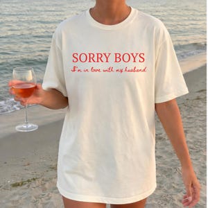 May include: Off-white t-shirt featuring the red text "SORRY BOYS" and "I'm in love with my husband." The shirt is worn on a beach, and a glass of rose wine is held in the hand.