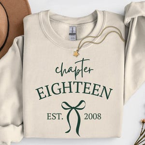 Può includere: Una felpa girocollo color crema con testo verde scuro che recita chapter EIGHTEEN EST. 2008 e una grafica centrale a fiocco. Una collana color oro con un ciondolo a forma di stella poggia sullo scollo del capo.