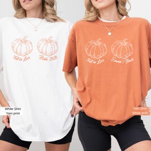 May include: A white t-shirt with orange pumpkin graphics and the text Fall in Love Bride 2026, and an orange t-shirt with white pumpkin graphics and the text Fall in Love Emma's Bach. Both shirts feature short sleeves and crew necklines.