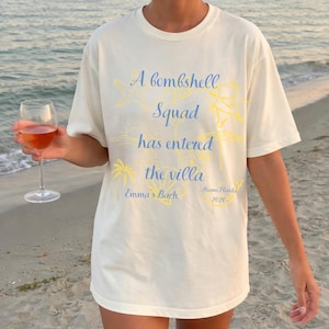May include: Off-white t-shirt featuring the text "A bombshell Squad has entered the villa" in blue and yellow. The shirt also includes "Emma's Bach" and "Miami Florida 2026". A person is holding a glass of rose wine on a beach.