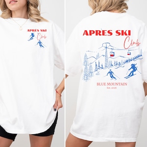 以下が含まれることがあります： 赤と青で「APRES SKI Club」とプリントされた白いTシャツ。前面にはスキーヤーが描かれ、背面にはスキーヤー、リフト、そして「BLUE MOUNTAIN Est. 2026」の文字が描かれた山の風景が描かれています。