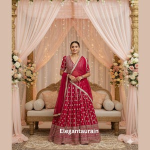 May include: A red embroidered lehenga with a matching dupatta. The lehenga features a detailed pattern and a decorative border. The outfit is worn by a person in front of a decorated backdrop with flowers and lights. The text "Elegantaurain" is visible.