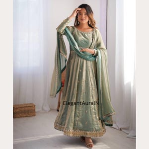 Peut inclure: Une robe Anarkali vert sauge avec un dupatta assorti. La robe à manches longues présente des broderies complexes et une jupe fluide. Le dupatta est de couleur sarcelle contrastante. Le texte "ElegantAuraIN" est visible sur la robe.