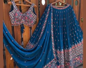 May include: A teal-colored, embroidered two-piece outfit. The top is a sleeveless crop top with intricate floral patterns in pink, red, and gold. The skirt is a long, pleated design with matching embroidery along the hemline. The outfit is displayed on wooden hangers.