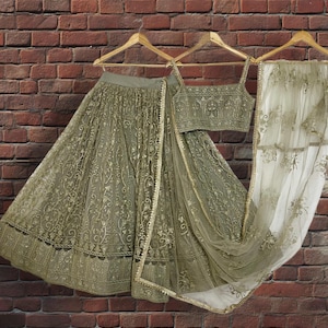 May include: An olive green embroidered lehenga choli set, including a skirt, top, and dupatta, hanging on wooden hangers against a brick wall. The set features intricate gold detailing and a sheer dupatta.