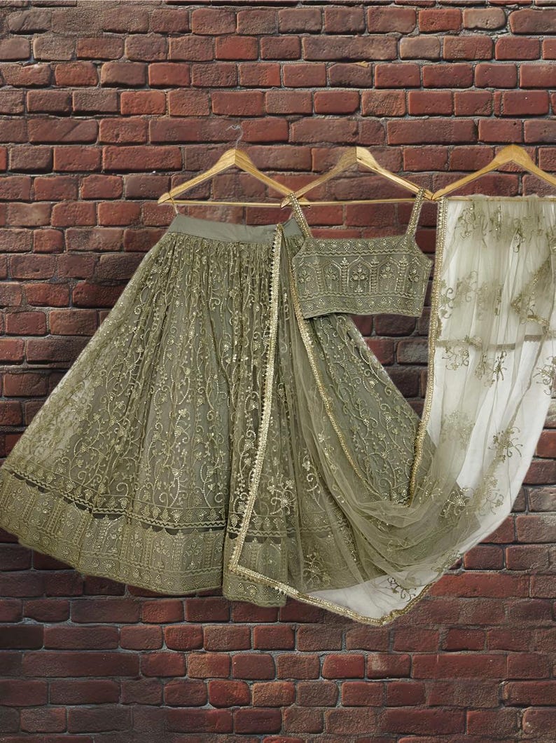 May include: An olive green embroidered lehenga choli set, including a skirt, top, and dupatta, hanging on wooden hangers against a brick wall. The set features intricate gold detailing and a sheer dupatta.