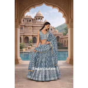 Könnte beinhalten: Ein grau-weiß gemustertes Lehenga Choli, ein traditionelles indisches Outfit, mit passendem Dupatta. Das Outfit weist aufwendige Stickereien und einen dekorativen Rand auf. Der Hintergrund zeigt ein verziertes Gebäude mit Bögen und einem Pool.