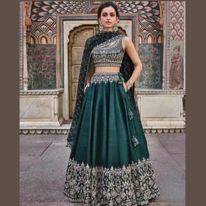 Puede incluir: Un lehenga esmeralda sin mangas con intrincados bordados dorados en la falda y el corpiño. El atuendo incluye un dupatta a juego y una parte superior detallada y adornada. El fondo presenta un entorno arquitectónico ornamentado.