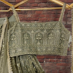 May include: Olive green embroidered crop top and skirt set with gold accents, hanging on a wooden hanger. The top features intricate floral and geometric patterns. The skirt has a matching design and is made of a sheer fabric.