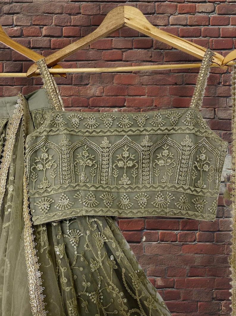 May include: Olive green embroidered crop top and skirt set with gold accents, hanging on a wooden hanger. The top features intricate floral and geometric patterns. The skirt has a matching design and is made of a sheer fabric.