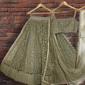 May include: An olive green lehenga choli set, including a skirt, top, and dupatta, hangs on a wooden hanger. The skirt and top are embellished with intricate gold embroidery. The dupatta has a gold border. The set is displayed against a brick wall.