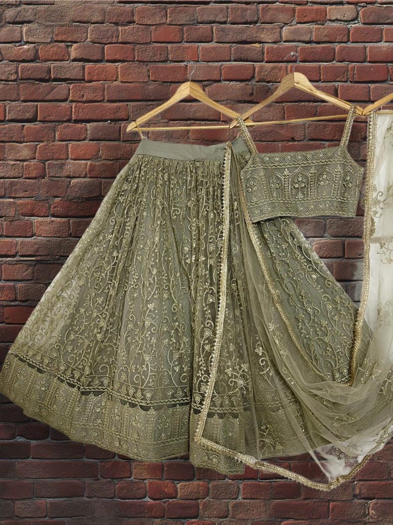 May include: An olive green lehenga choli set, including a skirt, top, and dupatta, hangs on a wooden hanger. The skirt and top are embellished with intricate gold embroidery. The dupatta has a gold border. The set is displayed against a brick wall.