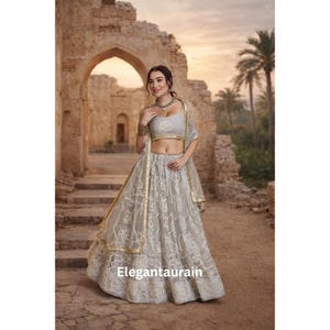 May include: A light gray lehenga with intricate lace detailing, a matching cropped top, and a sheer dupatta with gold trim. The outfit is worn by a person standing in front of an ancient stone structure. The text "Elegantaurain" is visible on the skirt.