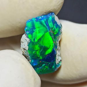 May include: A vibrant, raw opal gemstone with a striking display of color. The stone features a mix of vivid green, deep blue, and hints of red, creating a mesmerizing play of light. The opal is set against a neutral background, highlighting its natural beauty.