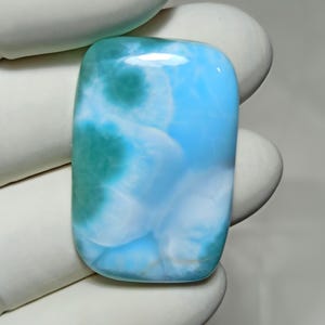 Puede incluir: Una gema rectangular pulida, con un color base azul claro y patrones arremolinados blancos y turquesas. La piedra tiene bordes redondeados y un acabado brillante, lo que sugiere que es un cabujón o similar.