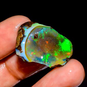 May include: A vibrant, raw opal gemstone with a mix of colors. The opal displays a striking play of color, with flashes of green, blue, and orange. The stone has a rough, natural texture and is held in a hand against a black background.