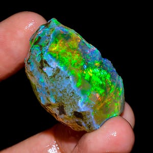 May include: A vibrant, rough-cut opal gemstone, showcasing a spectrum of colors. The stone displays vivid green, blue, and yellow hues, with a textured surface. The opal is held in a hand, highlighting its natural, raw form. The gemstone is set against a black background.