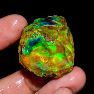 May include: A rough, uncut opal gemstone with vibrant colors. The opal displays a mix of green, yellow, and blue hues, with a textured surface. The gemstone is held in a hand against a black background.