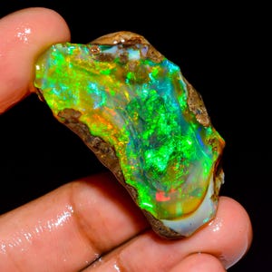May include: A vibrant, raw opal gemstone with a striking display of color. The opal features a mix of green, yellow, and blue hues, with flashes of red. The stone is held in a hand, showcasing its natural, rough texture.