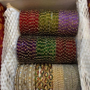 May include: A collection of stacked bangles arranged in three rows within a box. The top two rows feature vibrant red, green, purple, and maroon glass bangles with gold-tone accents. The bottom row includes metallic gold, silver, and teardrop-shaped stone-embellished bangles.