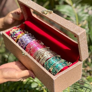 Puede incluir: Un joyero rectangular de color champán brillante con tapa abatible y cierre dorado, que contiene una fila de brazaletes apilados en varios colores, incluyendo turquesa, plata, rosa, morado y rojo, todos con pequeños cristales incrustados.