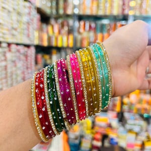Op de afbeelding: Een verzameling kleurrijke armbanden in rood, groen, roze, geel en turquoise. Elke armband is versierd met kleine, sprankelende strass-steentjes en goudkleurige randen. De armbanden zijn op een pols gestapeld en tonen hun levendige kleuren.