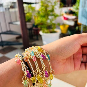 May include: A collection of gold-toned bangles adorned with colorful, dangling beads. The bracelets feature a delicate design with various shades of pink, yellow, green, and purple beads, creating a vibrant and eye-catching accessory.