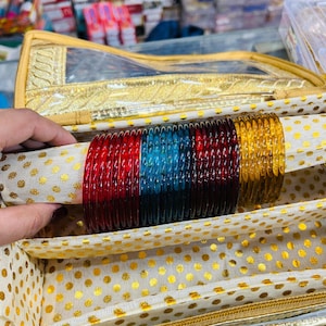 May include: A collection of colorful glass bangles in red, blue, and gold, displayed inside a gold-trimmed case with a cream-colored interior and gold polka dots. The bangles are arranged on a cream-colored fabric.