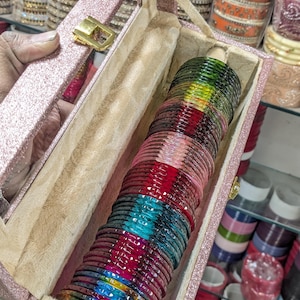 May include: A selection of vibrant glass bangles presented in a pink glitter box with a gold clasp. The bangles display a spectrum of colours including green, yellow, blue, pink, and red. Additional bangles are visible in the background.