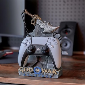 Può includere: Un controller grigio e nero per PlayStation 5 poggia su un supporto decorativo. Il supporto presenta il logo "God of War Ragnarok" e un design scolpito con una spada. Il controller è esposto su una superficie di legno.