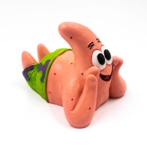 Op de afbeelding: Een roze, zeestervormig beeldje van Patrick Star uit de animatieserie Spongebob Squarepants. Het beeldje ligt, met een groene en paarse zwembroek. Het beeldje heeft zwarte ogen en een lachende mond.