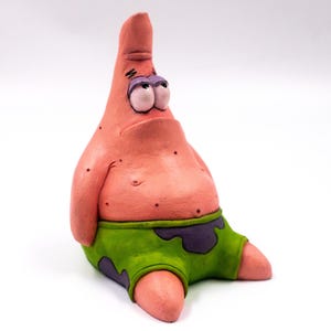 Op de afbeelding: Een roze, cartoonachtig beeldje van Patrick Star uit Spongebob Squarepants. Het beeldje heeft een verdrietige uitdrukking, een groene korte broek met paarse vlekken en zit. Het figuur heeft een lichtroze kleur met donkerroze sproeten.