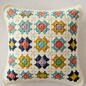 Puede incluir: Un cojín cuadrado de crochet color crema con un diseño de patchwork colorido. Cada cuadrado presenta un motivo floral central en tonos coral, azul, amarillo y morado, rodeado por un borde color crema. El cojín tiene un borde color crema.