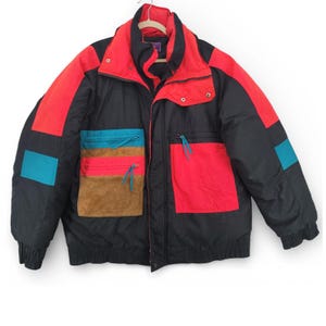 May include: A vintage black, red, and teal winter jacket. The jacket features a high collar, multiple pockets with teal and brown accents, and a red and black color block design. The jacket is hanging on a wooden hanger.
