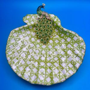 May include: A decorative seashell dish featuring a white base with a green lattice pattern and a gold-toned peacock brooch adorned with green and clear crystals resting on the upper edge of the shell, creating a detailed and textured display piece.