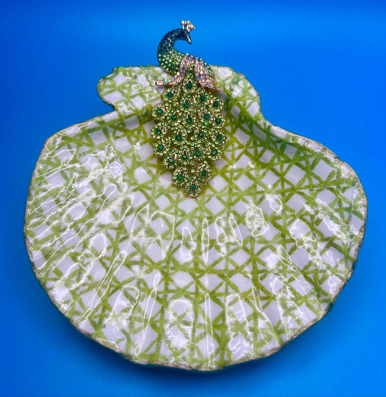 May include: A decorative seashell dish featuring a white base with a green lattice pattern and a gold-toned peacock brooch adorned with green and clear crystals resting on the upper edge of the shell, creating a detailed and textured display piece.