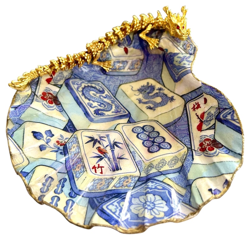 K&ouml;nnte beinhalten: Eine dekorative, muschelf&ouml;rmige Schale mit blau-wei&szlig;em Muster, das Mahjong-Steine zeigt. Die Schale ist mit Gold verziert und hat einen goldenen Drachenakzent. Die Steine zeigen Drachen, Bambus und andere traditionelle Symbole.