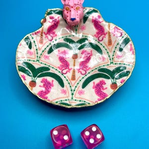 Op de afbeelding: Een decoratieve schelpvormige schaal met een geschilderd patroon van roze luipaarden en groene palmbomen op een cr&egrave;mekleurige achtergrond, bekroond met een roze luipaardkop en goudkleurige randen, vergezeld door twee transparante roze dobbelstenen.