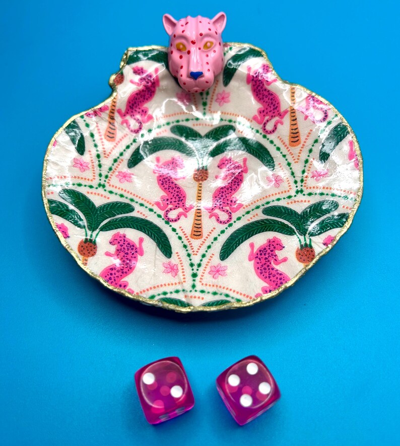 Op de afbeelding: Een decoratieve schelpvormige schaal met een geschilderd patroon van roze luipaarden en groene palmbomen op een cr&egrave;mekleurige achtergrond, bekroond met een roze luipaardkop en goudkleurige randen, vergezeld door twee transparante roze dobbelstenen.