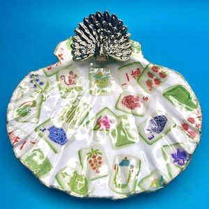 May include: A white scallop shell trinket dish featuring a decoupage pattern of various Mahjong tiles with colorful illustrations. A metallic bronze-toned peacock figurine with an open fan tail is attached to the top edge of the shell.
