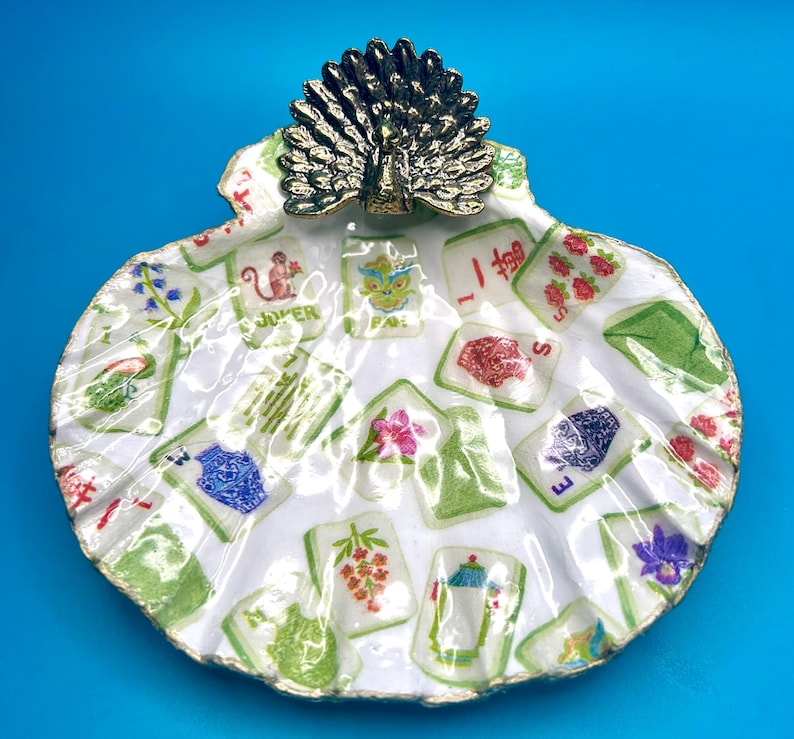 May include: A white scallop shell trinket dish featuring a decoupage pattern of various Mahjong tiles with colorful illustrations. A metallic bronze-toned peacock figurine with an open fan tail is attached to the top edge of the shell.