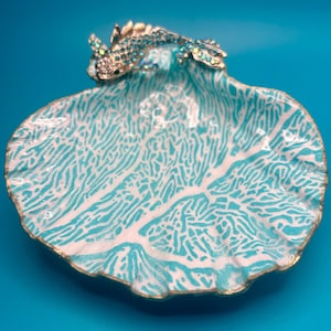 May include: A decorative shell-shaped dish featuring a white and turquoise coral-like pattern on the interior surface. A metallic fish ornament adorned with small, sparkling light blue crystals is attached to the top edge of the shell dish.