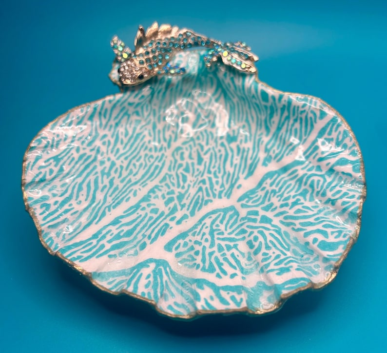 May include: A decorative shell-shaped dish featuring a white and turquoise coral-like pattern on the interior surface. A metallic fish ornament adorned with small, sparkling light blue crystals is attached to the top edge of the shell dish.