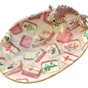 May include: An oval-shaped decorative dish with a gold rim, featuring a pink and white patterned interior with illustrations of rectangular tiles. A jeweled fish figurine with pink and clear crystals is attached to the dish.
