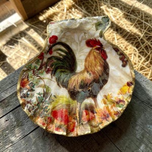 May include: A decorative scallop shell dish featuring a decoupage design of a rooster with brown, green, and red feathers. The shell is decorated with colorful floral patterns in shades of red, yellow, and green, with a gold-painted scalloped edge.