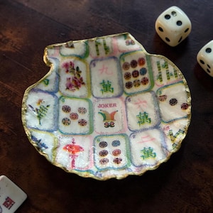 May include: A decorative, shell-shaped dish featuring a colorful Mahjong tile design with a gold-painted rim. The dish includes the word "JOKER" and is accompanied by two white dice with black dots. The dish is on a dark wooden surface.