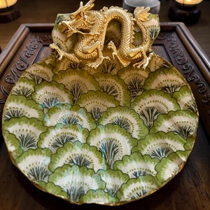 May include: A decorative shell-shaped dish featuring a gold-colored dragon figurine attached to the top edge. The interior surface of the dish is decorated with a repeating pattern of green and white fan-shaped motifs with dark green line details.