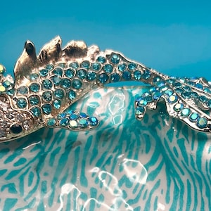 May include: A silver-toned metal fish brooch featuring a body encrusted with shimmering light blue rhinestones and a head accented with clear crystals and a single black eye, positioned over a white and turquoise patterned ceramic dish.