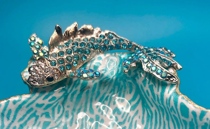 May include: A silver-toned metal fish brooch featuring a body encrusted with shimmering light blue rhinestones and a head accented with clear crystals and a single black eye, positioned over a white and turquoise patterned ceramic dish.